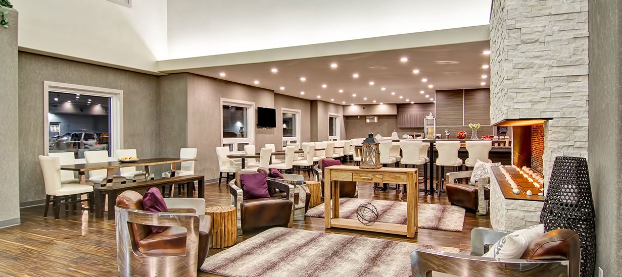 A comprehensive view of the breakfast and lounging areas of d3h Home Inn & Suites South Saskatoon which showcases the towering white limestone electric fireplace surrounded by leather club chairs and a beige rustic console table adorned with art objects.  The breakfast area is populated by eating tables and benches and a sea of white upholstered Parsons dining chairs.