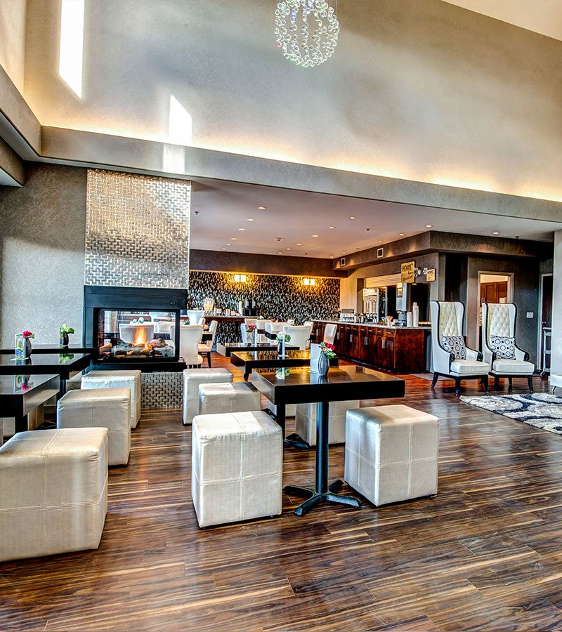 A double sided electric fireplace, set in a metallic silver herringbone pattern surround, provides warmth to the breakfast and lounge area of d3h Home Inn & Suites Swift Current.  Dark wood square dining tables matched with white cube shaped pouf seating and upholstered dining chairs are placed throughout the eating space.  The elongated mahogany wood breakfast bar holds countertop appliances, eating utensils and dinnerware.