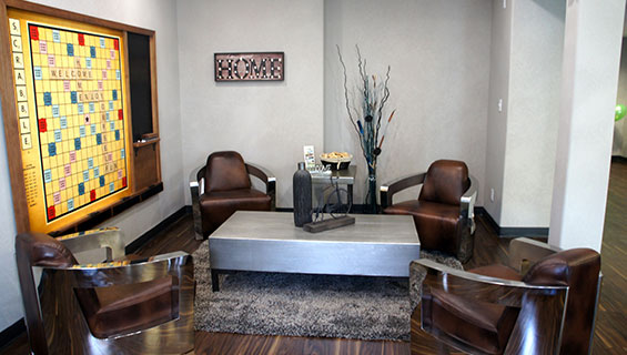 Four leather club chairs encircle a heavy steel top rectangular coffeetable placed over an area rug in the guest lounge area at Home Inn Express in Medicine Hat, Alberta.  A large scrabble board hangs over a white-gray wall, with an attached black chalkboard for keeping score. 