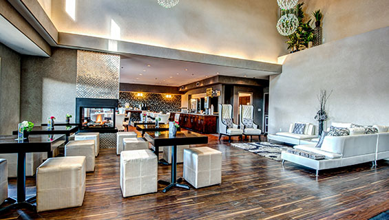 A panoramic view of the lounging and eating areas of the d3h Home Inn & Suites Yorkton:  The gleaming metallic herringbone pattern surround of the electric fireplace stands out amidst a sea of eating tables and white seating contrasted against a black and white graphical accent wall at the back of the dining area.  The lounge space showcases a white angular sectional sofa paired up with two white high backed chairs embellished with black arm trim.