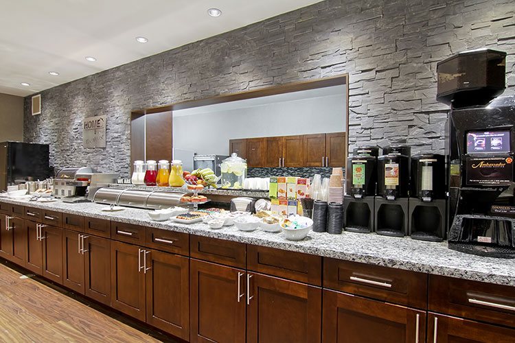 The HomeEssentials breakfast area, offering complimentary breakfast for guests staying at d3h Home Inn & Suites hotels, features a long granite top breakfast bar bearing, pitchers of juice, fruit, baked goods, and amenities such as countertop food warmers, beverage dispensers, and toasters.