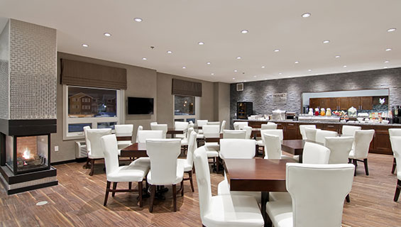 A panoramic view of the HomeEssentials breakfast area at d3h Home Inn & Suites Regina Airport showcases a long granite countertop stocked with food and appliances placed up against a gray stone back wall with a cut out view of the kitchen.  Square chocolate brown eating tables and white upholstered dining chairs are placed throughout the area, with a glass enclosed gas fireplace in the corner.