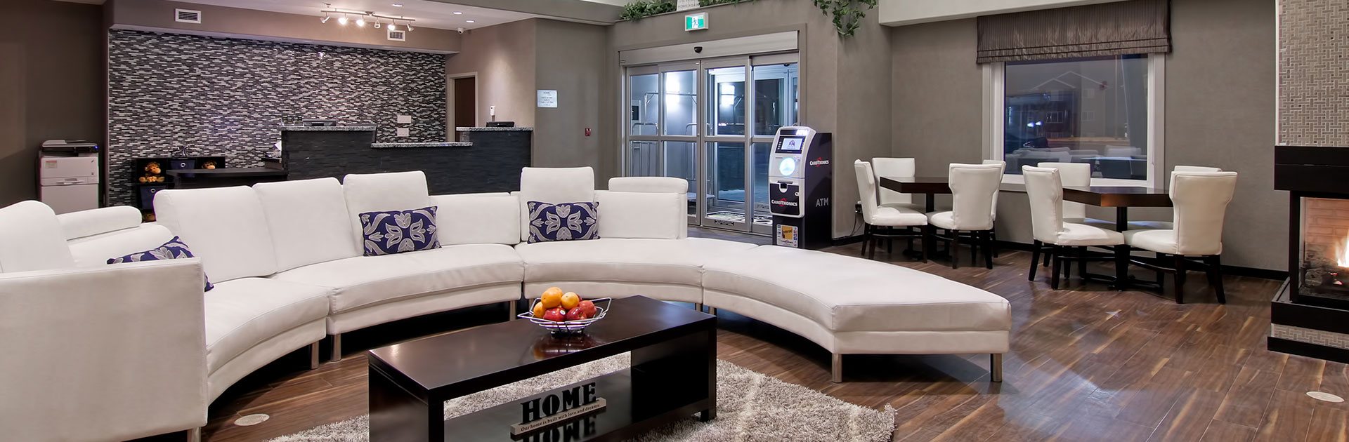 The lobby of the d3h Home Inn & Suites Regina Airport features a bright white curved sectional sofa with decorative purple accent pillows contrasted against the black and white pattern accent wall and charcoal gray stone check in counter in the background.