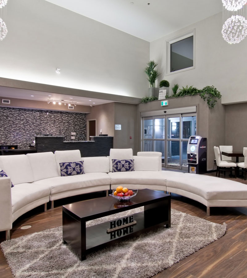 The lobby of the d3h Home Inn & Suites Regina Airport hotel showcases a semi-circular white sectional sofa decorated with white and purple accent pillows and a solid dark wood coffeetable placed over an area rug.  An ATM machine is placed by the glass door entrances while in the background, stands the dark stone check-in counter.