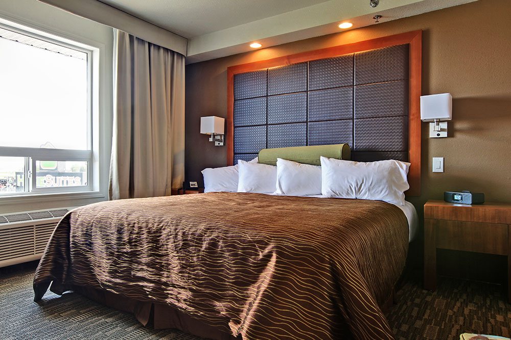 The King Suite at d3h HomeSuites Regina East showcases a king sized bed dressed in layers of white pillows, a pale green bolster pillow and copper brown and gold print comforter.  The dark leather upholstered headboard is contrasted against the coffee colored walls, which boasts lamps with cube shaped shades mounted on both sides of the bed.