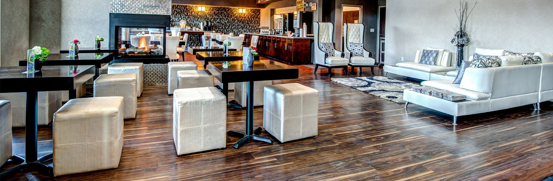 Black brown square eating tables with white cube shaped pouf ottoman seating are placed around a burning electric fireplace inset with black trim at the breakfast room at d3h Home Inn & Suites in Yorkton, Saskatchewan.