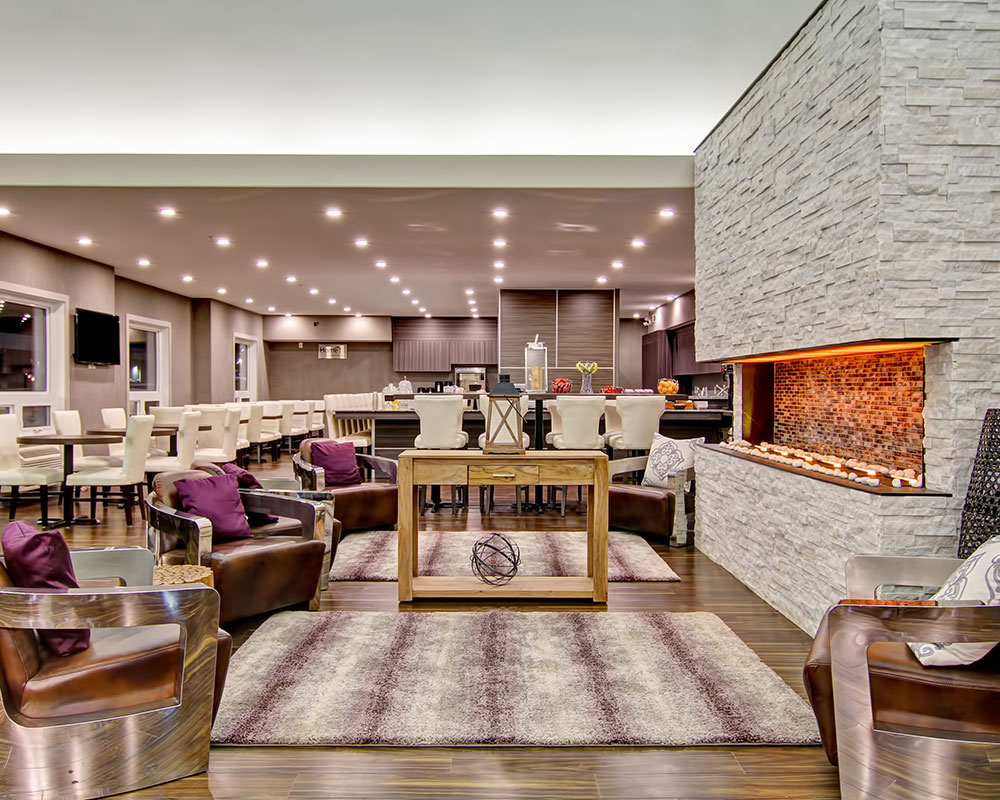 The large white birch limestone electric fireplace provides heating to the lounging and dining area of d3h Home Inn & Suites Saskatoon South.  Leather club chairs with thick metal arms surround the fireplace and a rustic console table.  The woodgrain laminate wall panels of the breakfast bar are seen in the background amidst rows of square eating tables and benches placed amongst a sea of carefully arranged white upholstered dining chairs.