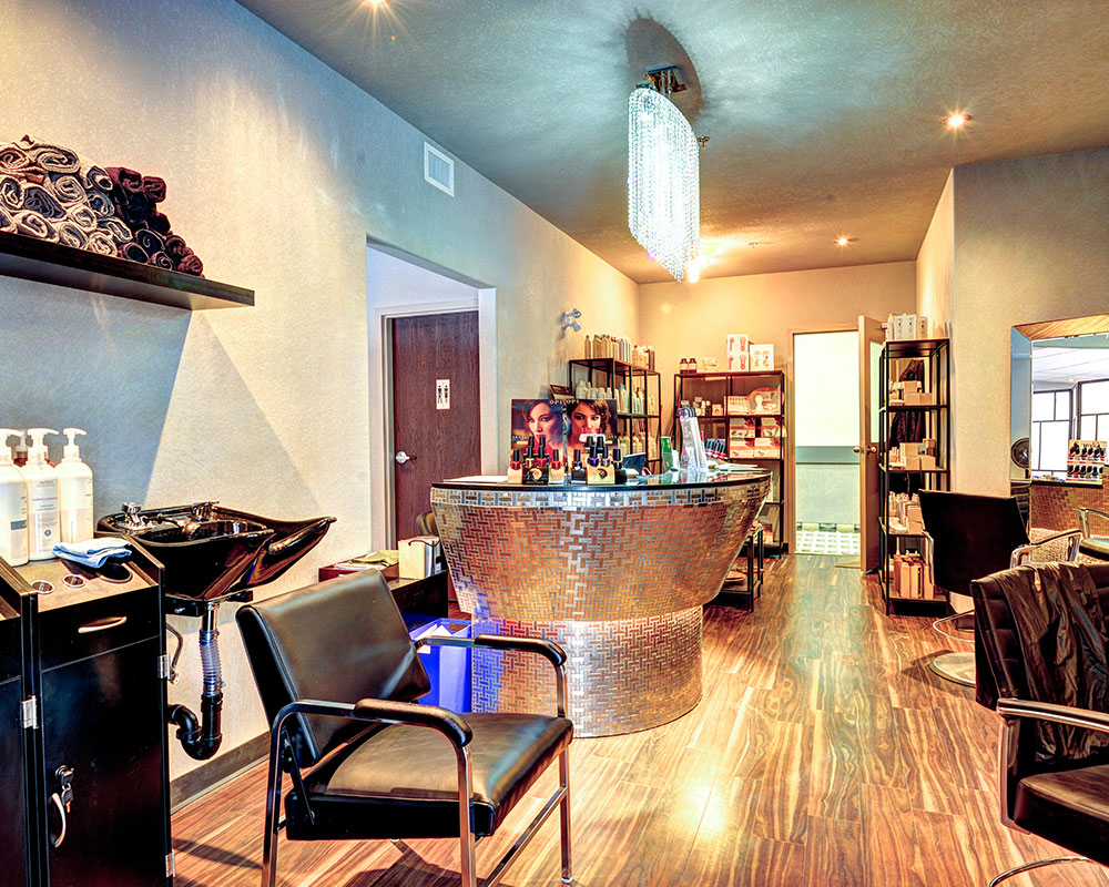 Spa facilities at a d3h managed property.  In the center of the space sits a round silver metallic herringbone patterned reception desk, displaying merchandise and pamphlets.  In the background, three multi-level black metal shelving units display more merchandise for sale, while in the foreground, a black vinyl reclining chair is tucked under a black shampooing basin sink.