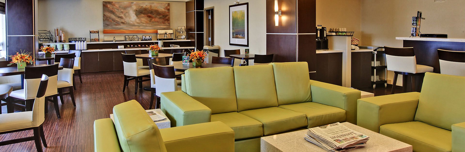 A modern furniture set is featured in the lounge space at d3h Home Inn & Suites Yorkton: a vinyl chartreuse green sofa and armchair set is matched to solid cube shaped coffee and end tables.  A coffee bar is set in a secluded corner next to the breakfast area, while a two-tiered elongated counter, bearing stainless steel appliances and eating ware, is lined up against the back wall of the dining area.  A series of square tables and white upholstered dining chairs are placed throughout the space.