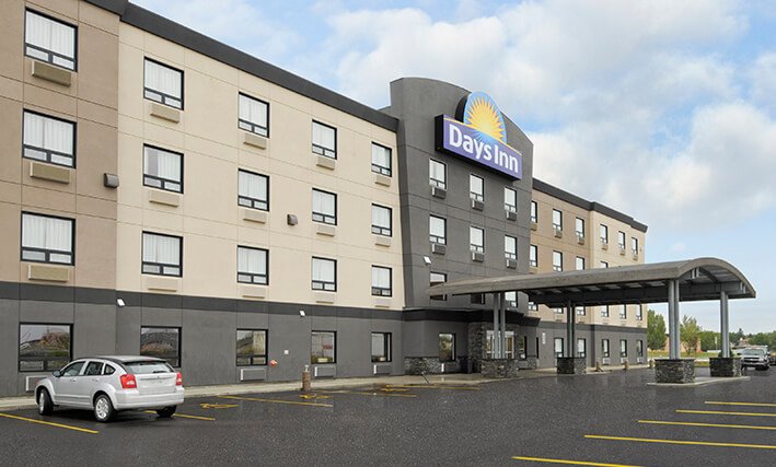 The parking lot view of the d3h Days Inn Regina Airport in Saskatchewan:  Two vehicles are parked in the vicinity of the semi-circular shaped portico over the glass entrance to the four storey hotel.  The hotel is tri-tone colored (painted beige, tan and seal-gray), with a large rendering of the corporate logo (brand name in white bold lettering and yellow sunrise icon against a blue background) above the portico-covered entrance.