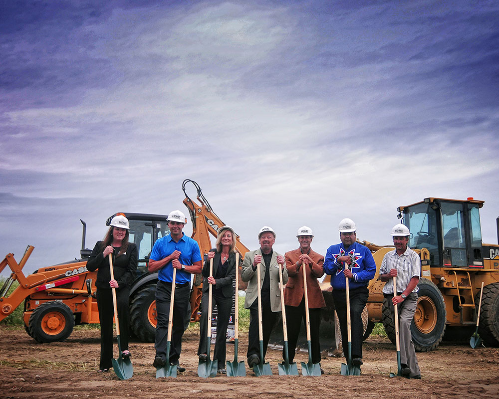 A group of two women and five men from the management team at d3h hotels, wearing construction hard hats, are all holding shovels dug into the dirt of a development site.  Two tractors are seen in the background.