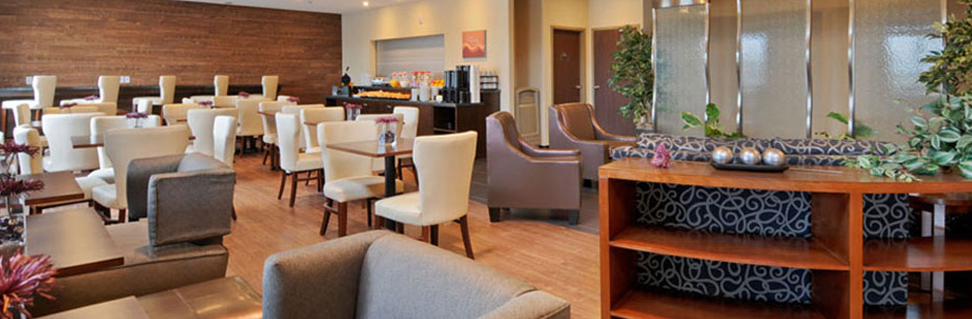 The Daybreak Café at d3h Days Inn Regina Airport West features a woodgrain accent back wall with built-in elongated eat-in bench and white upholstered seating.  The breakfast counter is fully stocked with fruit, baked goods and beverages, while rows of square tables and white upholstered dining chairs are placed throughout the eating area.  The adjacent lounge area is furnished with a pair of peanut brown vinyl armchairs and swirl patterned navy blue loveseat.