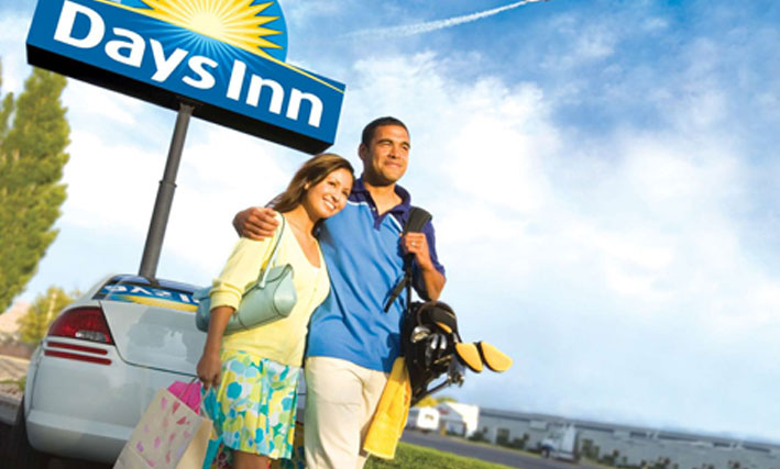 A photo of a happy young couple, the female carrying shopping bags and the male with golf clubs slung over his shoulder, stand before a parked white car and the Days Inn corporate sign (represented by a bright yellow sunrise icon against a blue background with the corporate name in white typeset), making their way to their booked suite.