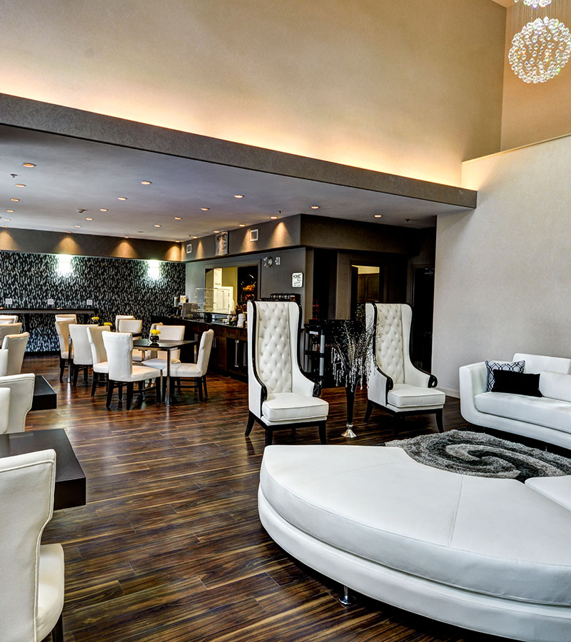 The HomeEssentials breakfast room at d3h Home Inn & Suites Swift Current is adorned with two white high back armchairs in the foreground, square dark brown wood eating tables surrounded by white upholstered dining chairs and a distinct black and white pattern accent wall in the background.